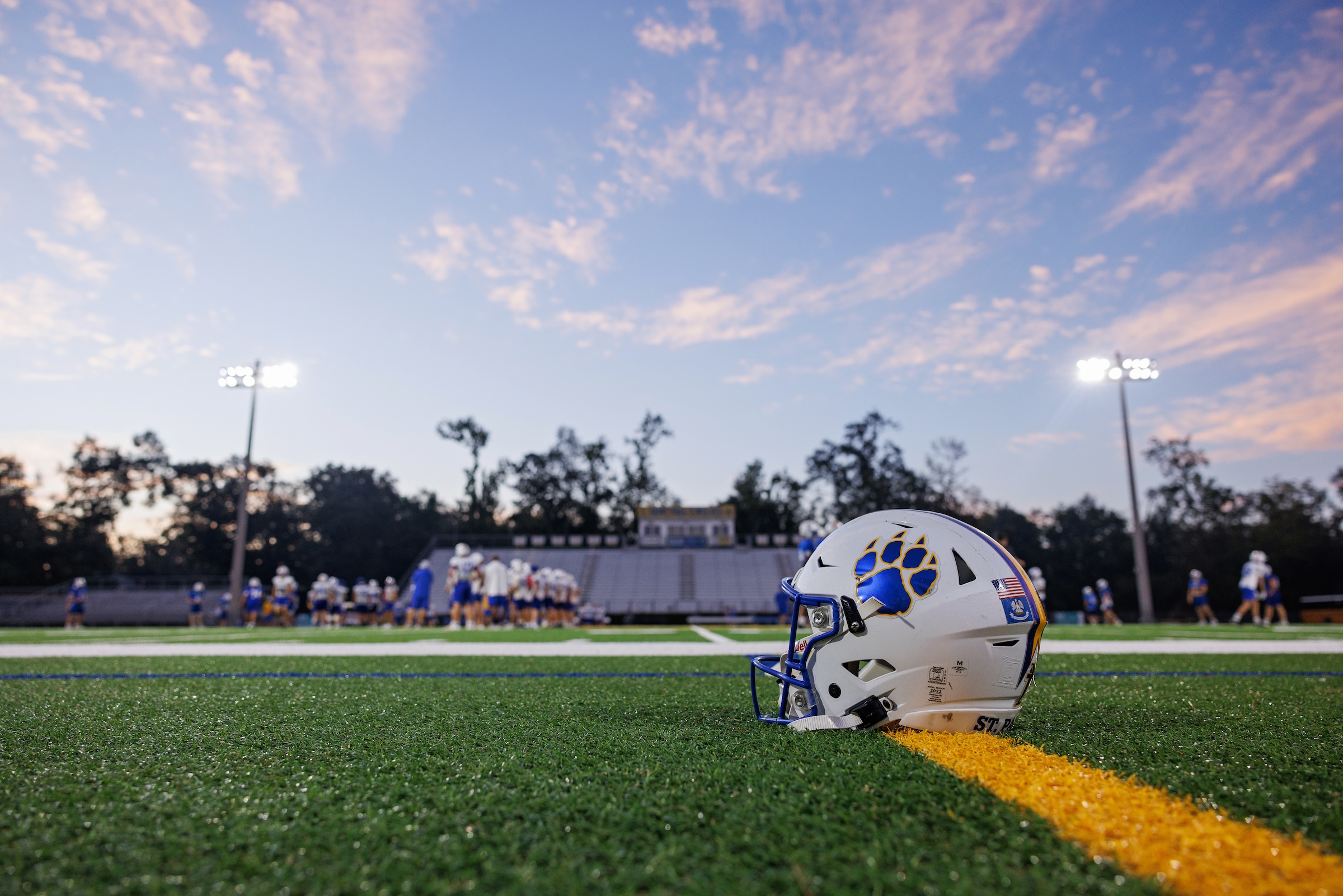 A white St. Paul's School football helmet with a blue paw print sits on green turf, players and stadium lights in the background.