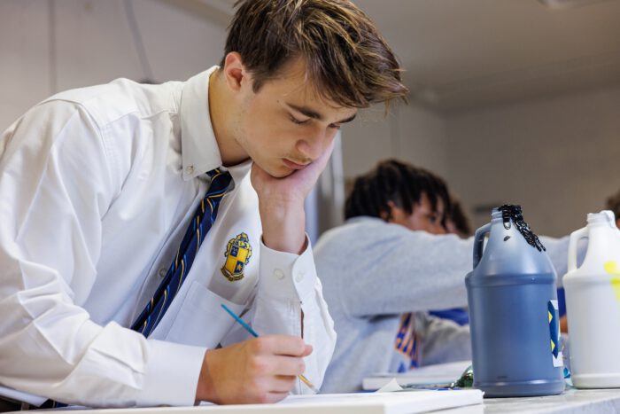 A student at St. Paul's School paints at his desk, resting his head on his hand. Art supplies scatter the table in this private school scene.