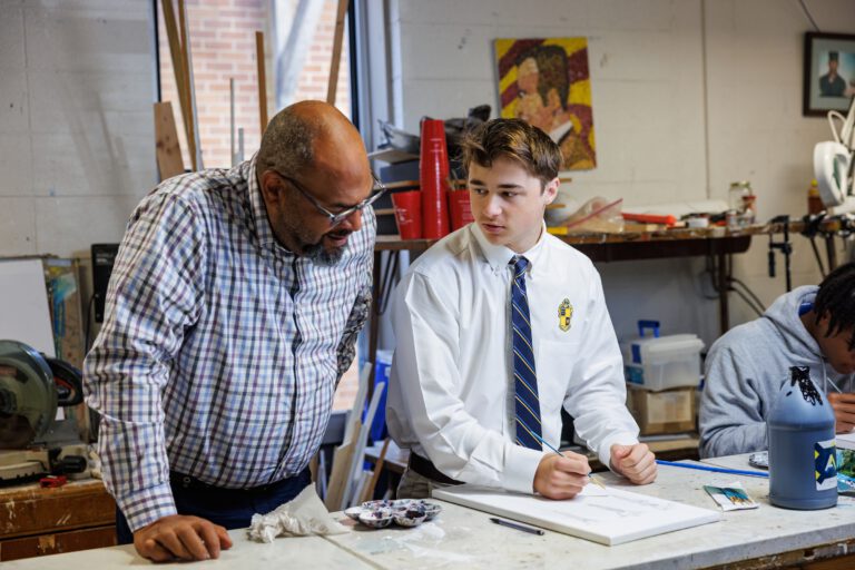 A teacher and student in uniform talk at a table in an art classroom at St. Paul’s School, with art supplies and paintings in the background.