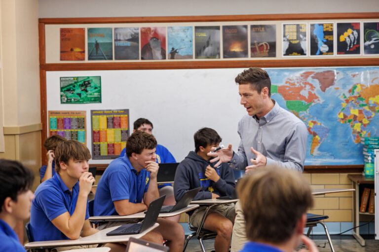 A teacher speaks to students in a classroom at St. Paul's School, a leading private school on the Northshore with desks, laptops, and a world map.
