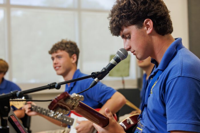 Two young men in blue shirts from St. Paul's School play electric guitars, with one singing into a microphone in a brightly lit room.