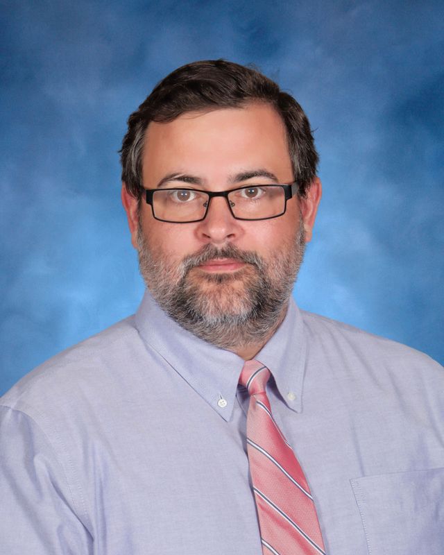 A man with short dark hair, glasses, and a beard in a light purple shirt and pink striped tie, representing St. Paul's School.