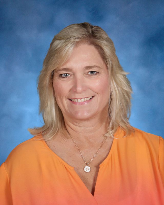 A woman with blonde hair in an orange top and necklace smiles, representing St. Paul's School against a blue studio background.
