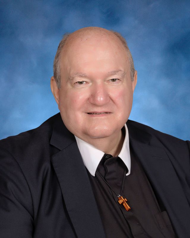 An older man in a dark suit, clerical collar, and cross necklace poses before a blue backdrop at St. Paul’s School, a school for boys on the Northshore.