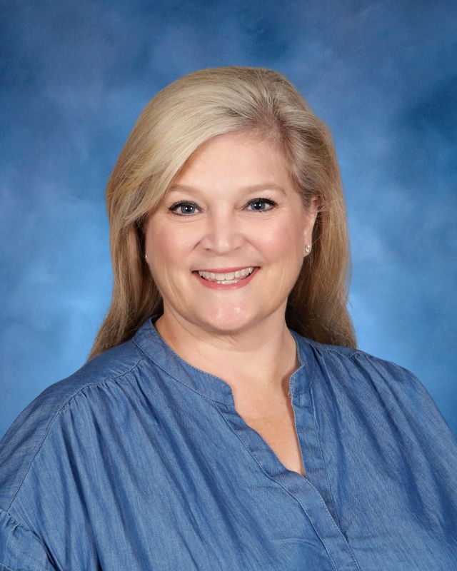 Smiling woman with long blonde hair in a blue blouse, posed before a blue background at St. Paul's School, a leading school for boys northshore LA.