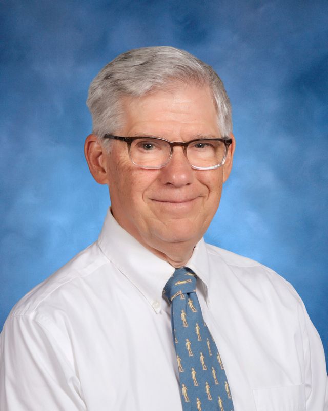 An older man with gray hair and glasses, in a white shirt and blue tie, poses before a blue backdrop at St. Paul's School on the Northshore.