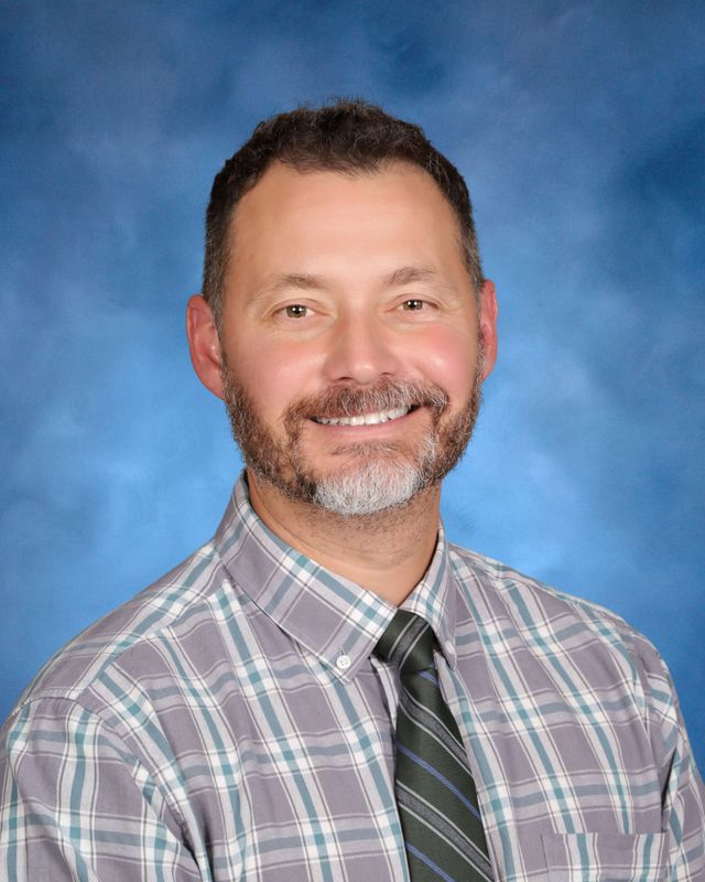 A man with short dark hair and a beard smiles in plaid and a tie, pictured at St. Paul's School, a top school for boys Northshore LA.