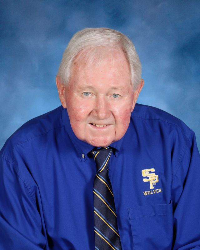 Older man with short white hair in a blue dress shirt and "SP Wolves" tie, posed before a blue background at St. Paul’s School.