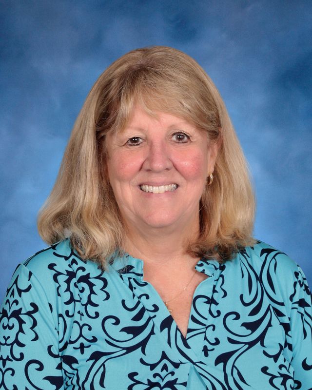 A woman with blonde hair in a blue patterned blouse smiles in front of a blue studio background at St. Paul's School.