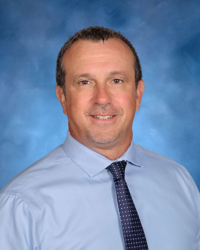 A man in a light blue dress shirt and polka dot tie poses against a blue background, reflecting the spirit of St. Paul's School.