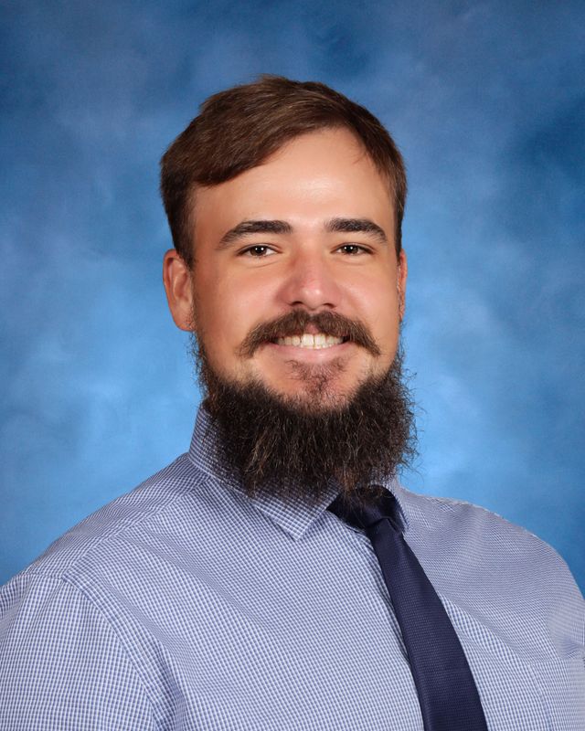 A man with brown hair and a full beard smiles, wearing a checkered shirt and tie, against a blue background at St. Paul's School.