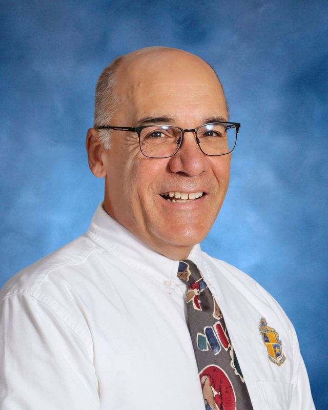 A middle-aged man with glasses smiles in a white shirt and colorful tie, proudly displaying the St. Paul’s School crest against a blue background.
