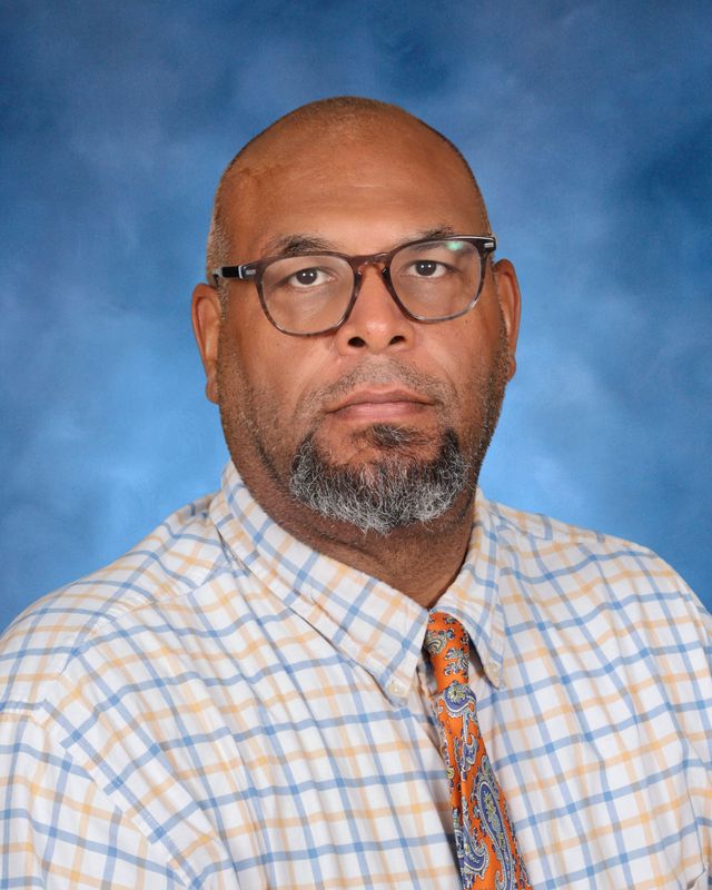 A man with glasses and a short beard, dressed in plaid and a tie, poses before a blue backdrop at St. Paul's School on the Northshore.