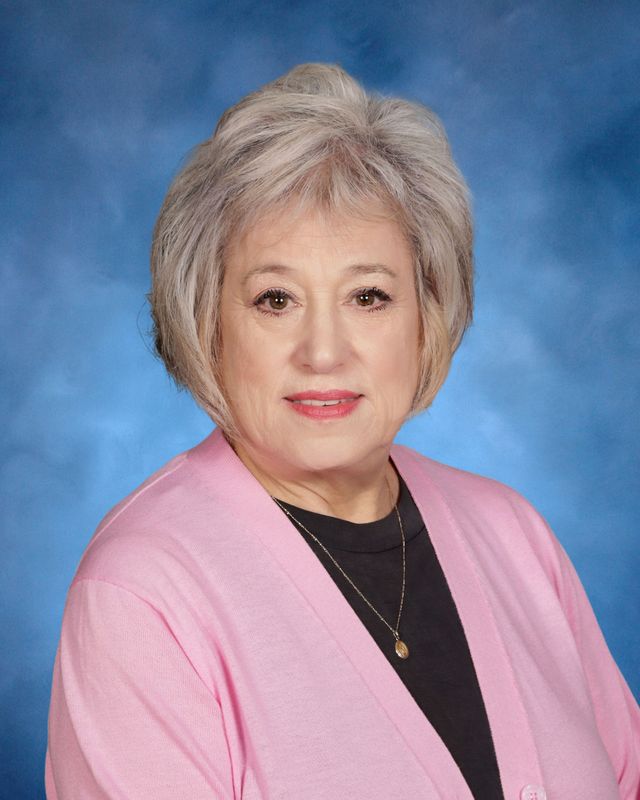An older woman with short gray hair in a pink cardigan poses before a blue studio background, reminiscent of portraits at St. Paul's School.