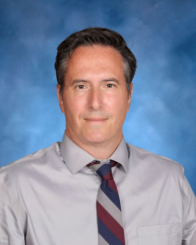 A man with short dark hair in a gray dress shirt and striped tie poses before a blue background at St. Paul's School, a school for boys Northshore LA.