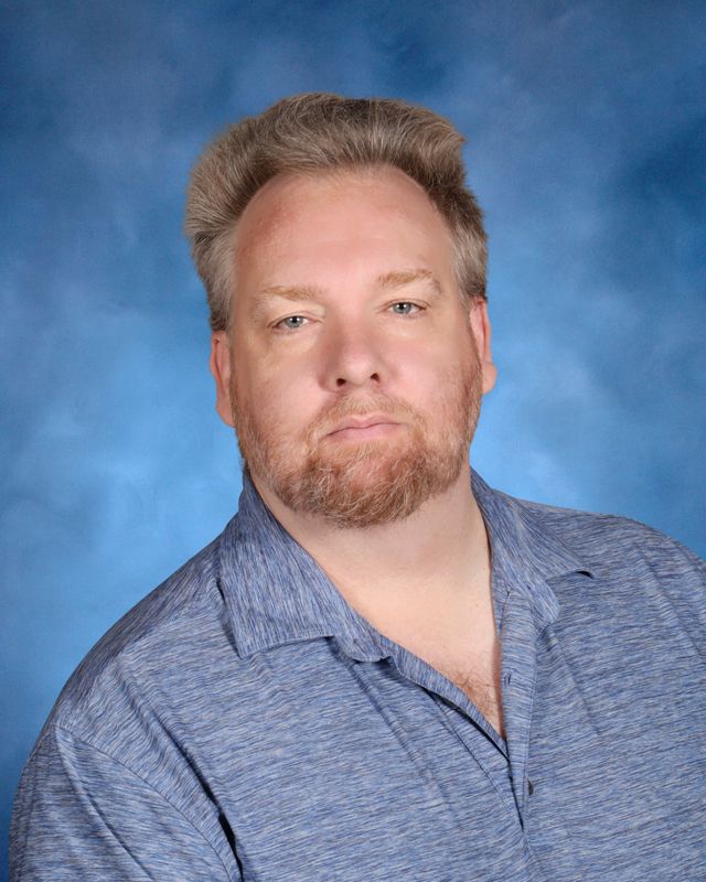 A man with light hair and a beard in a blue shirt poses before a blue studio backdrop at St. Paul’s School, a leading private school on the Northshore.