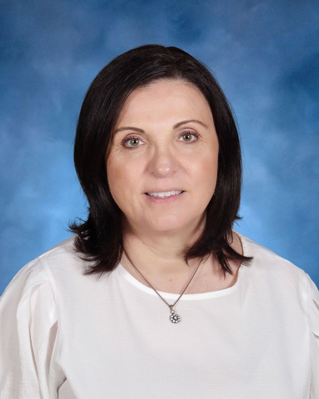 Woman with shoulder-length dark hair wearing a white top and pendant necklace, posing in front of a blue studio background at St. Paul’s School.