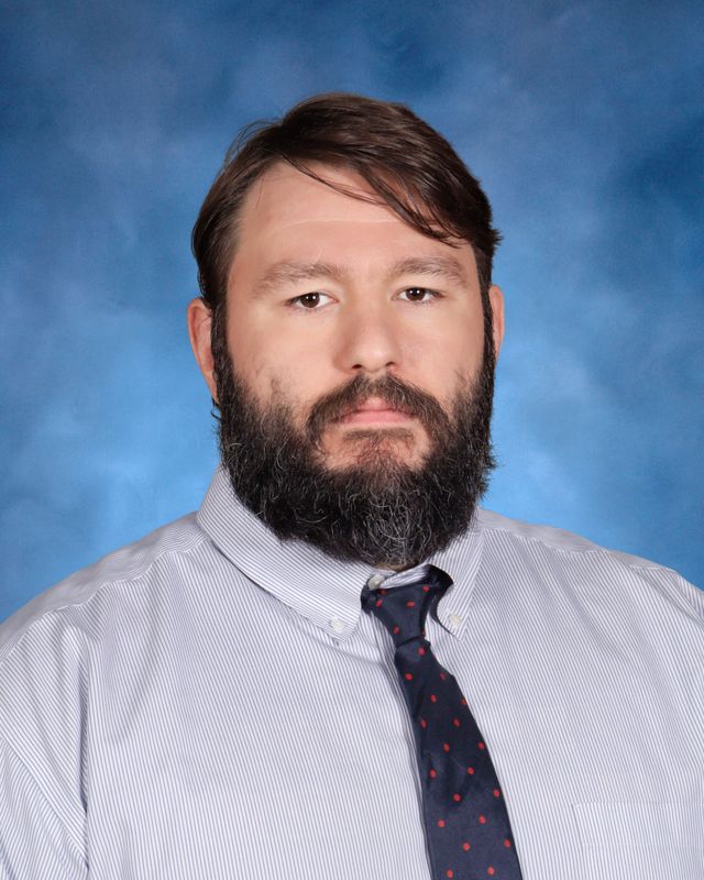 A man with a beard and mustache in a striped shirt and polka dot tie poses, representing St. Paul's School, a top school for boys on the Northshore.