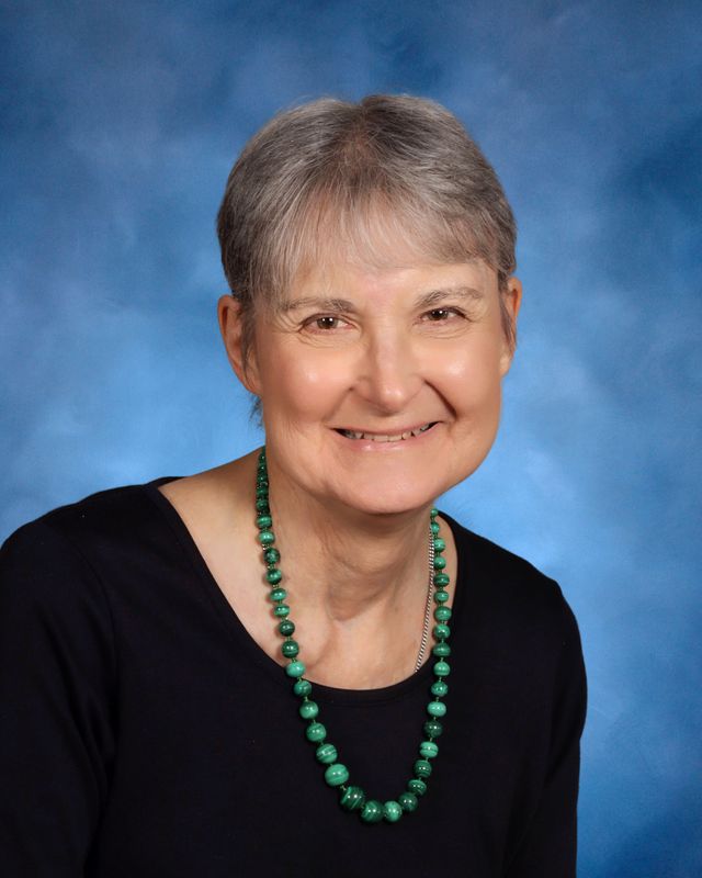 An older woman with short gray hair and a green beaded necklace smiles in front of a blue studio backdrop at St. Paul's School.