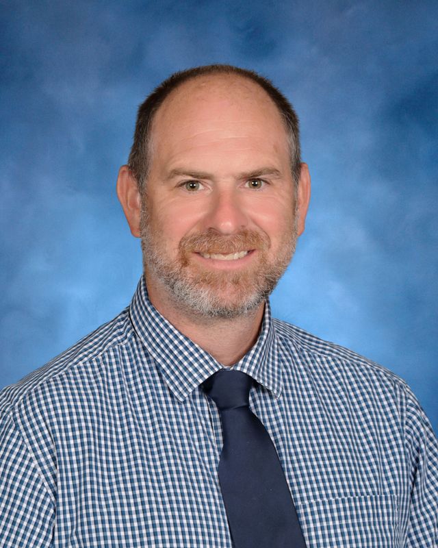A man with short hair and a beard, in a checkered shirt and tie, poses before a blue backdrop at St. Paul’s School on the Northshore.
