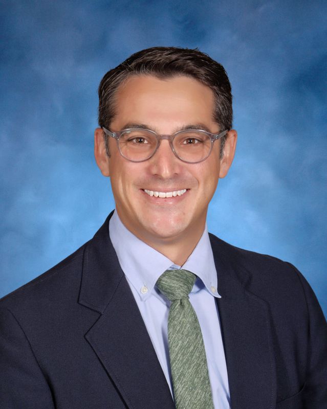 A man in a suit and glasses smiles before a blue studio background, representing St. Paul's School, a leading private school on the Northshore.