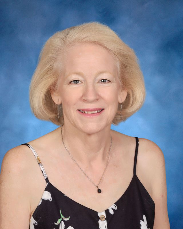 A woman with light blonde hair in a black floral dress and necklace poses before a blue studio backdrop, representing St. Paul's School.