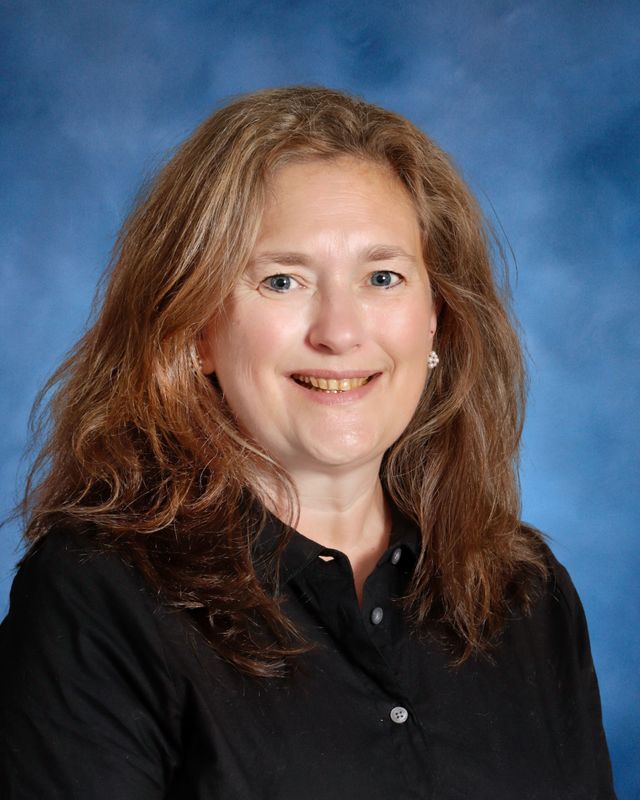 Woman with long light brown hair in a black button-up shirt smiles before a blue background at St. Paul's School, a private school on the Northshore.