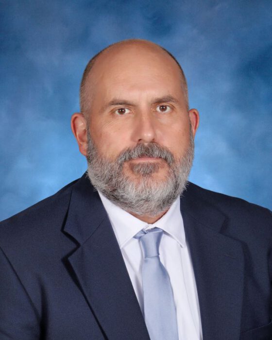 A man with a gray beard in a navy suit poses before a blue backdrop, representing St. Paul's School, a leading school for boys Northshore LA.