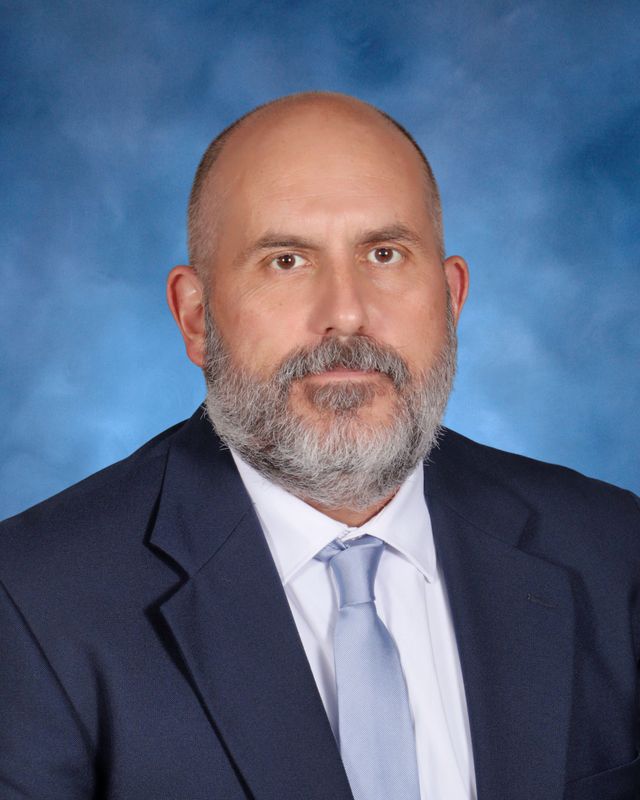A man with a gray beard in a navy suit poses before a blue backdrop, representing St. Paul's School, a leading school for boys Northshore LA.