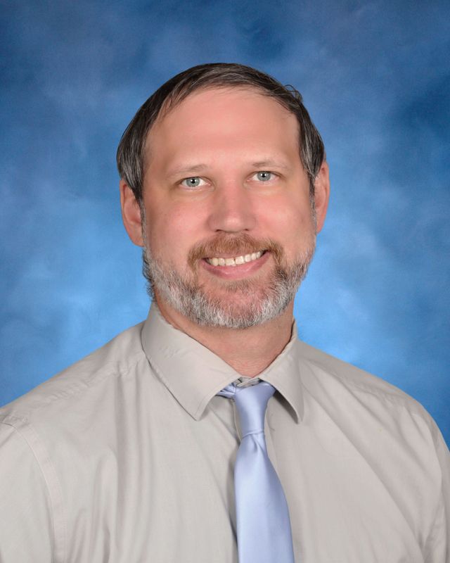 A man with short brown hair and a beard, in a light gray shirt and tie, poses before a blue background at St. Paul's School.