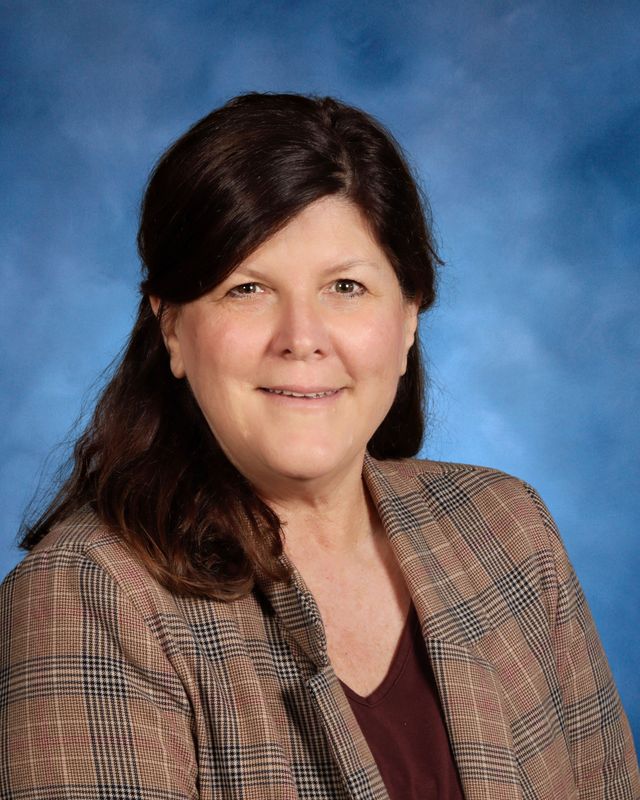 A woman with long brown hair in a plaid blazer and maroon top poses before a blue background, similar to portraits at St. Paul’s School.