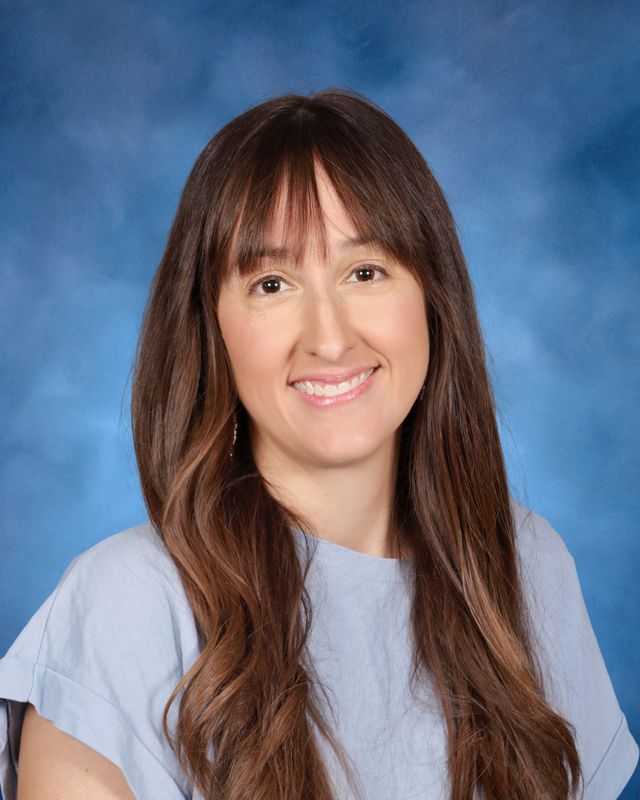 A woman with long brown hair and bangs smiles at the camera, representing St. Paul's School against a blue studio background.
