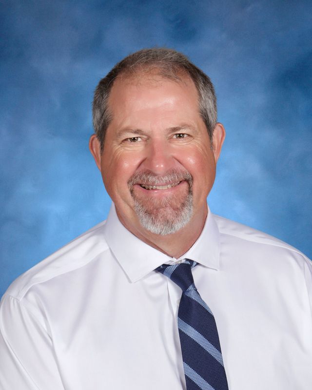 A middle-aged man with short gray hair and a goatee, wearing a white shirt and striped tie, smiles at St. Paul's School in front of a blue backdrop.