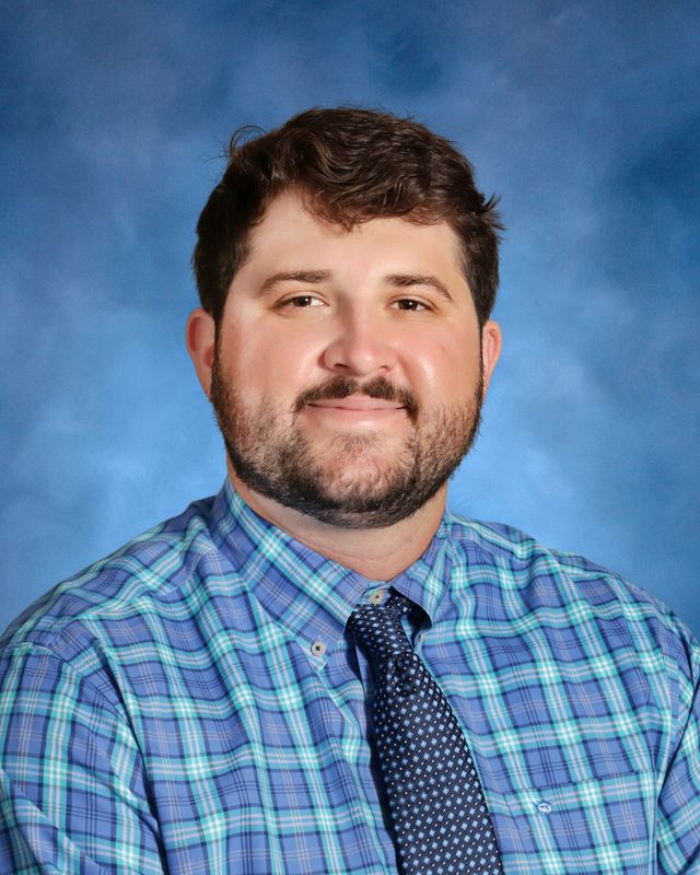 A man with short brown hair and a beard, in a blue plaid shirt and tie, poses against a blue studio backdrop at St. Paul’s School.