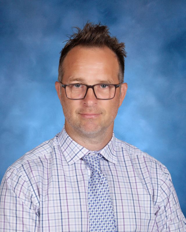 A man with short brown hair and glasses in a plaid shirt and tie poses before a blue backdrop at St. Paul’s School, a school for boys Northshore LA.