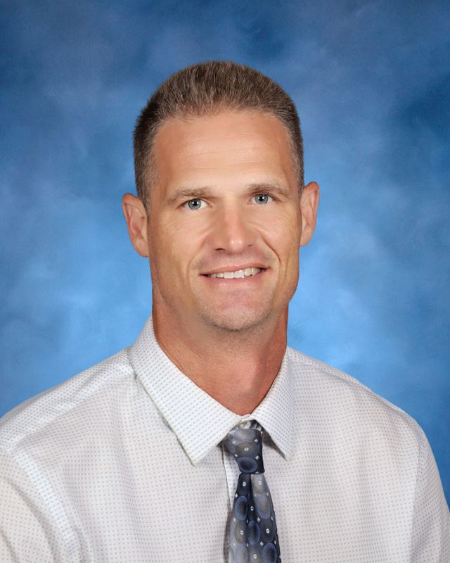 A man with short light brown hair, in a white shirt and tie, poses before a blue studio backdrop at St. Paul's School on the Northshore.
