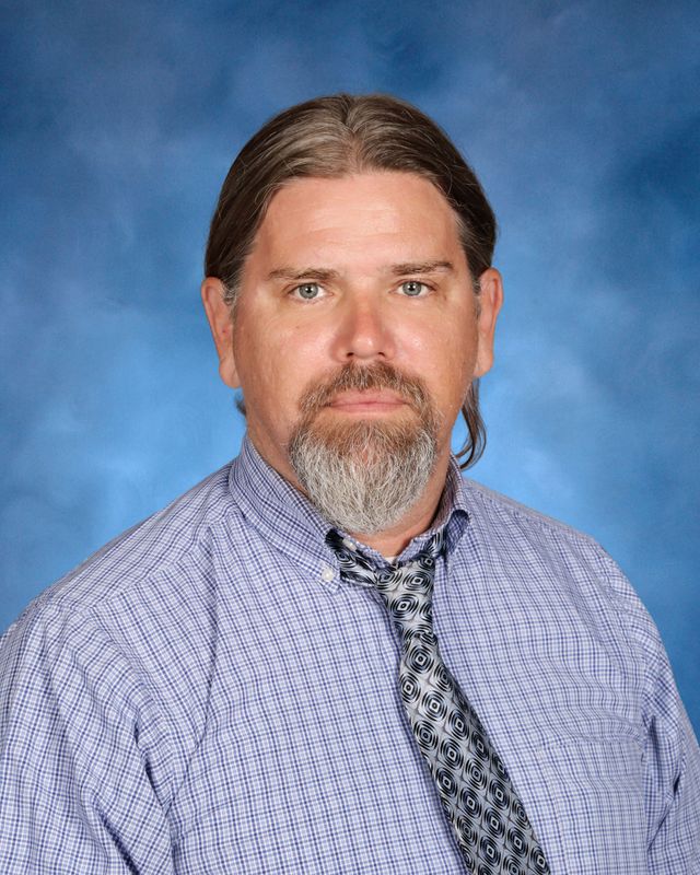 A man with long hair and a beard, in a checked shirt and tie, poses before a blue backdrop at St. Paul’s School on the Northshore.
