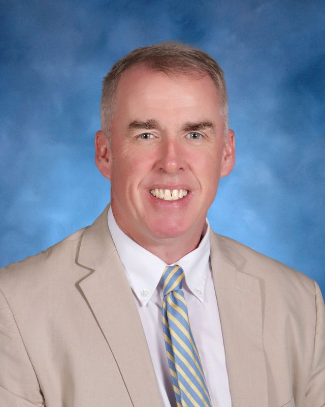 A man in a beige suit poses, smiling, in front of a blue backdrop at St. Paul's School, a leading private school on the Northshore.