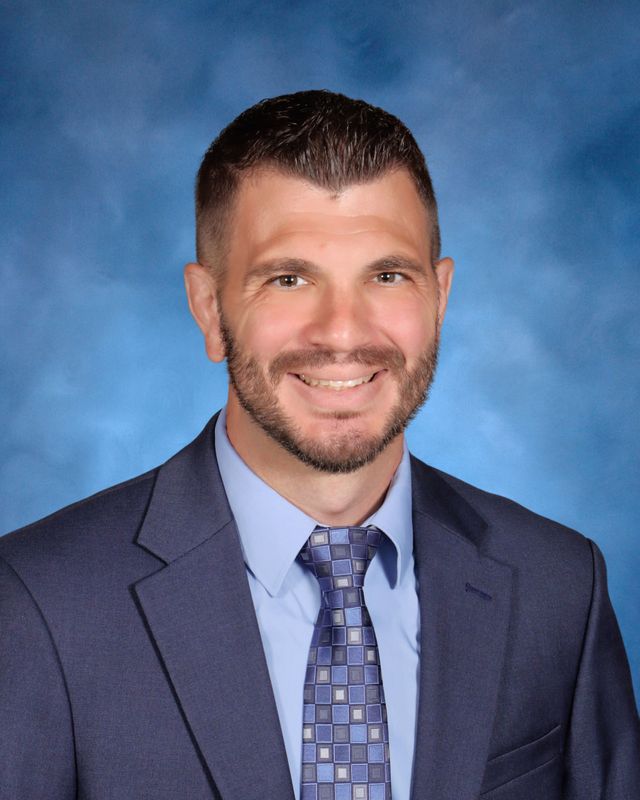 A man in a blue suit and tie smiles before a blue background, representing St. Paul's School, a top school for boys on the Northshore, LA.