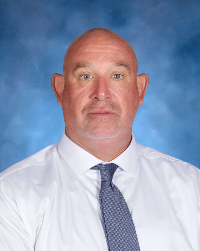 A bald man with light skin in a white shirt and blue tie poses before a blue background at St. Paul's School, a school for boys Northshore LA.