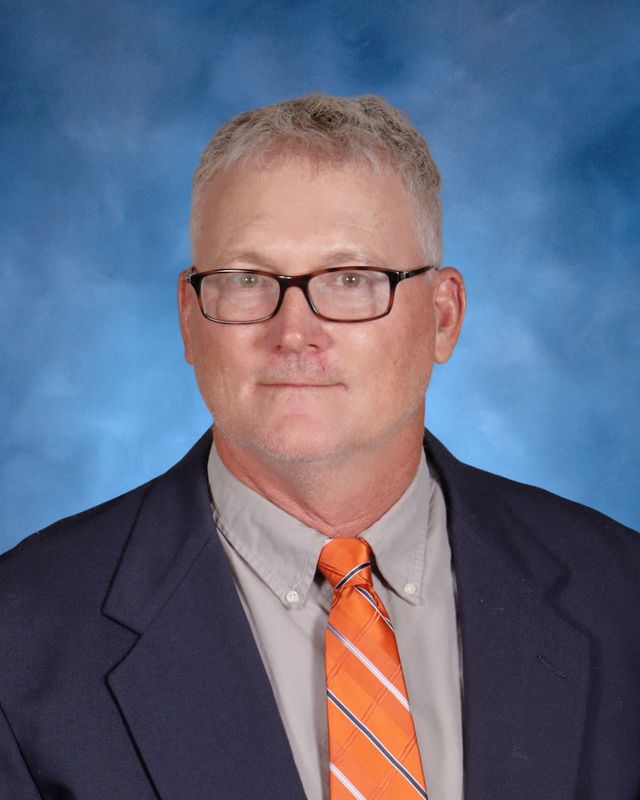 A middle-aged man with gray hair and glasses, in a navy suit and orange striped tie, poses before a blue backdrop at St. Paul's School.
