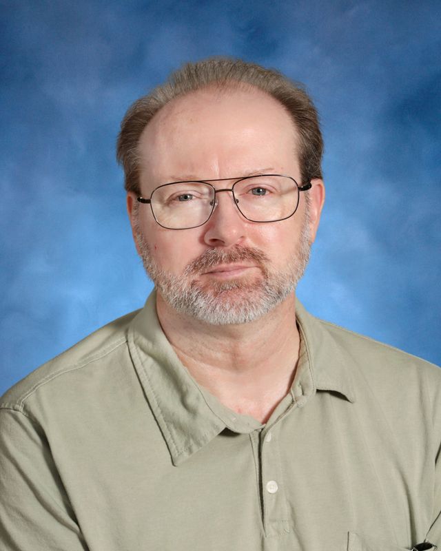 A middle-aged man with glasses, short hair, and a beard in a light green collared shirt represents St. Paul's School against a blue background.