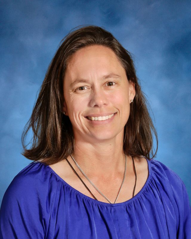 A woman with straight brown hair in a purple blouse smiles in front of a blue background, representing St. Paul's School on the Northshore.