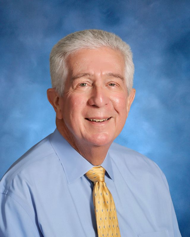 Older man with short gray hair wearing a light blue shirt and yellow patterned tie, representing St. Paul's School, in front of a blue background.