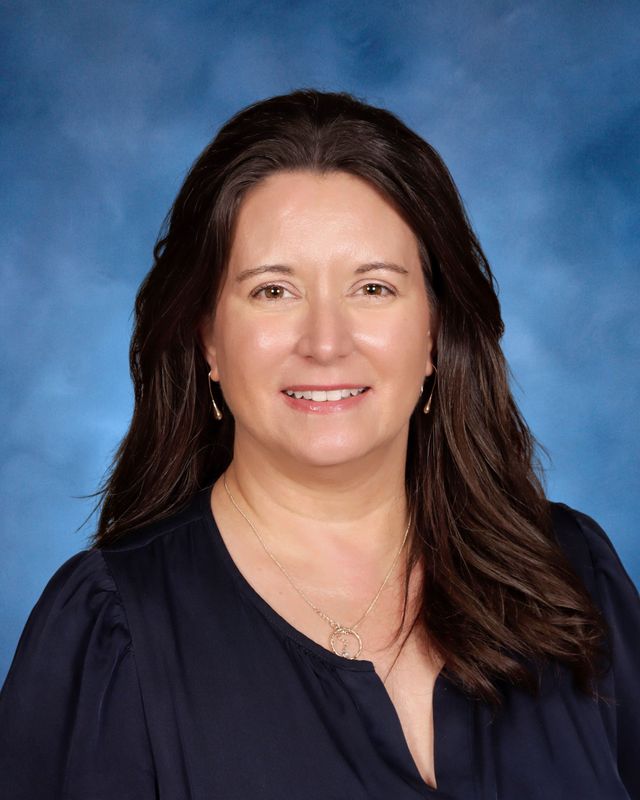 Woman with long brown hair, navy blouse, gold necklace, smiling in front of a blue studio background at St. Paul's School.