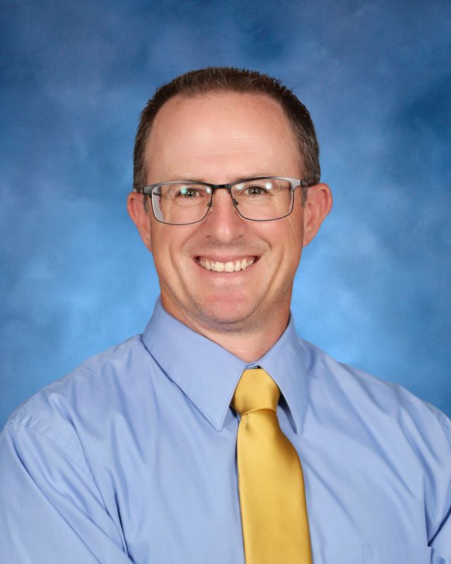 A man in glasses, a light blue dress shirt, and yellow tie smiles at the camera, representing St. Paul’s School for boys on the Northshore.