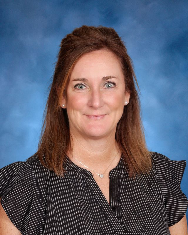 A woman with straight brown hair, in a black blouse with white stripes, poses before a blue background at St. Paul's School on the Northshore.