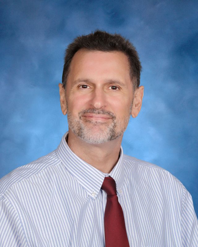 A man with short dark hair and a beard, in a striped shirt and red tie, poses against blue—representing St. Paul’s School on the Northshore.