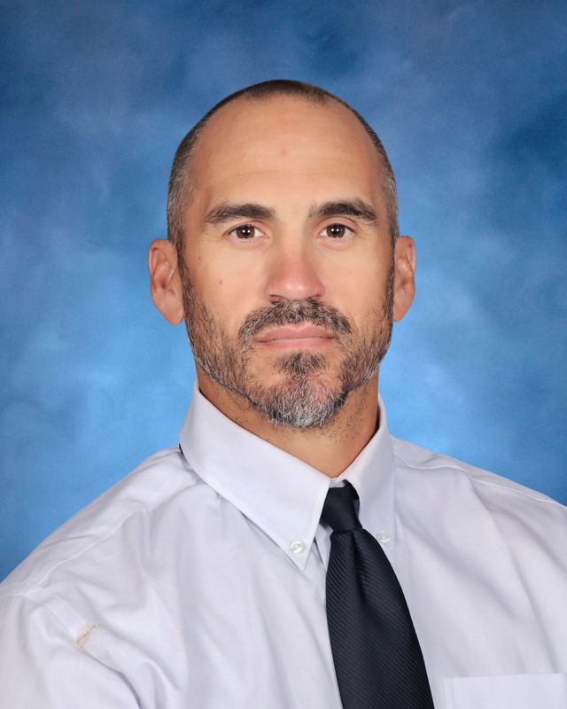 A man with short hair and a beard in a white dress shirt and black tie poses in front of a blue St. Paul's School background.