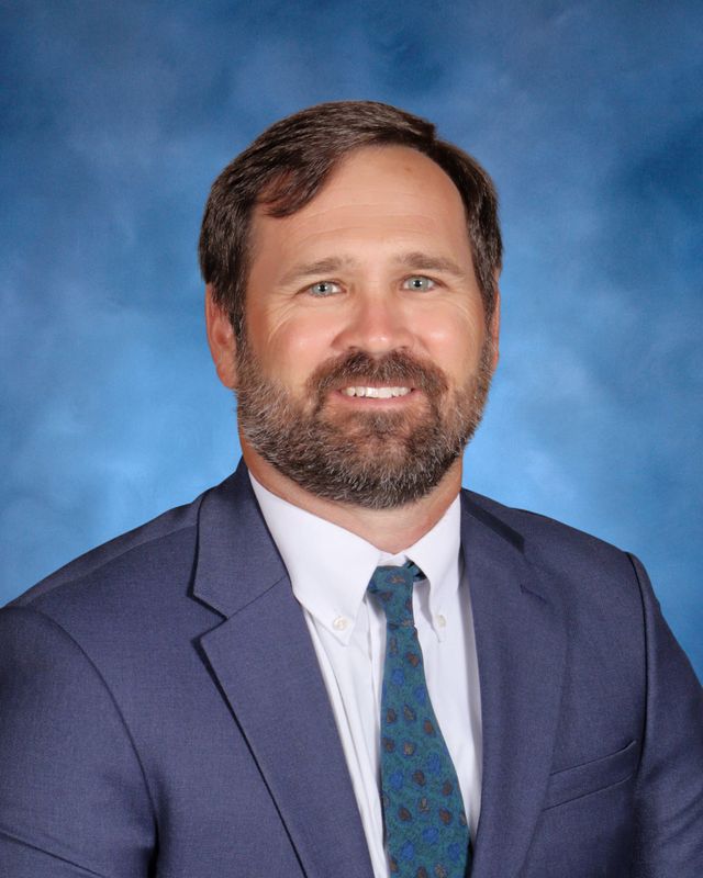 A man with brown hair and a beard in a blue suit, posed before a blue backdrop at St. Paul's School, a private school on the Northshore.